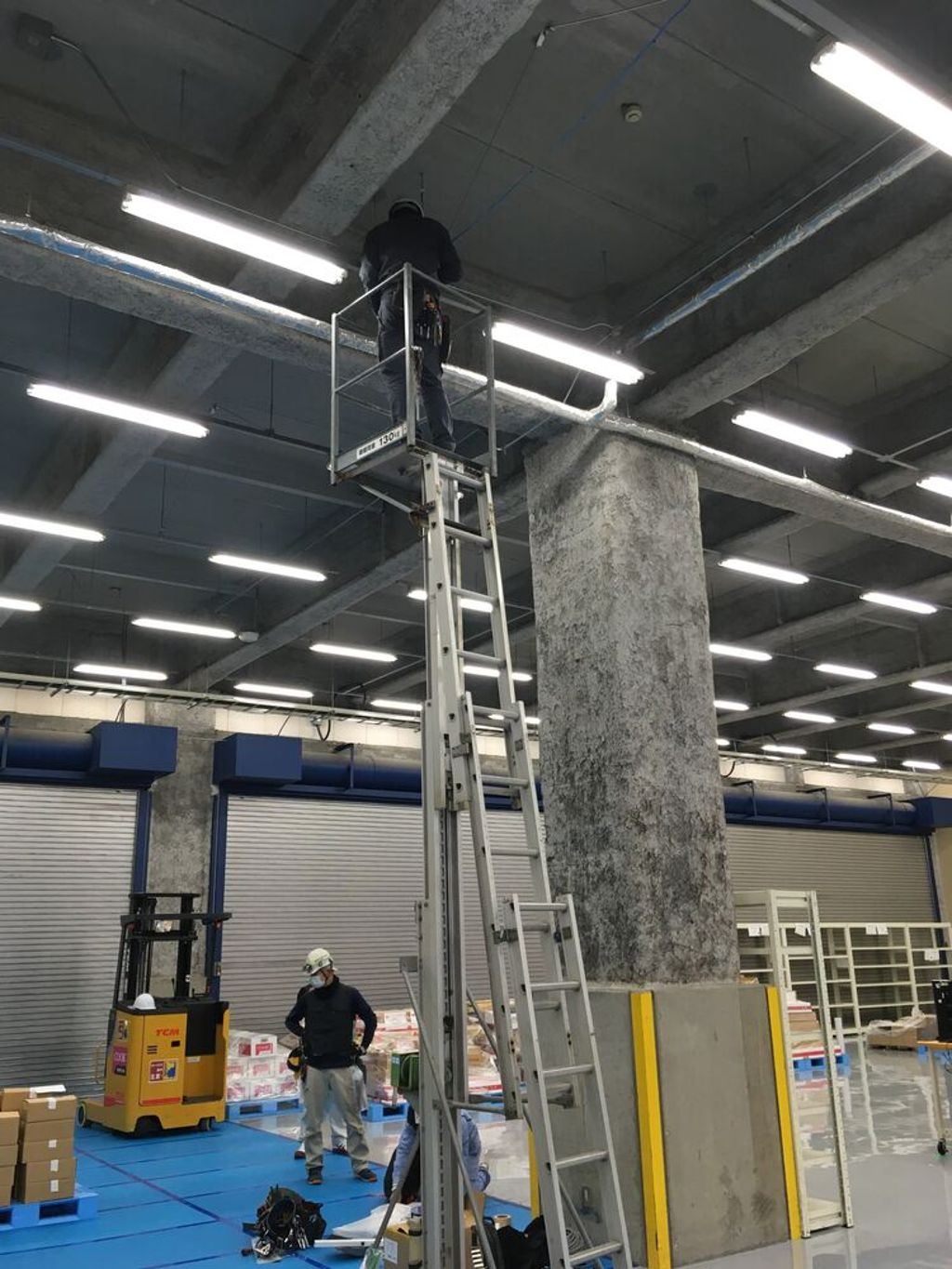 An eSolia technician on a tall platform ladder installs cabling at the ceiling of a warehouse while a colleague and a reach forklift work on the floor below